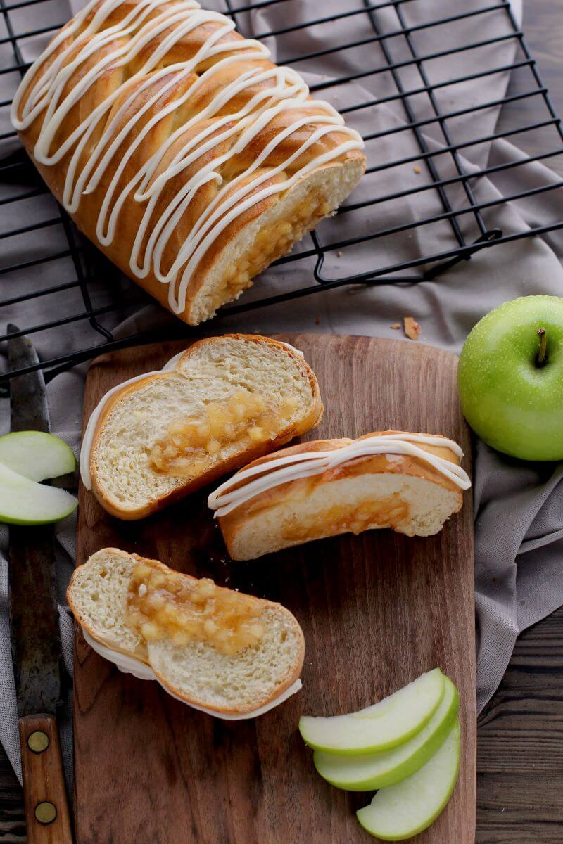 A partial Apple Braided Bread on a cooling rack. In front of the rack is a cutting board with three slices of the bread on it. The full bread and slices are iced. There's also a full green apple and several apple slices around the slices of bread.