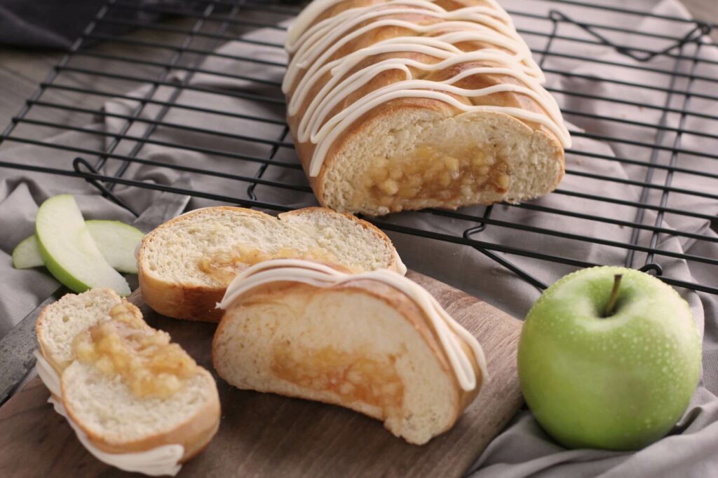 A partial Apple Braided Bread on a cooling rack. In front of the rack is a cutting board with three slices of the bread on it. The full bread and slices are iced. There's also a full green apple and several apple slices around the slices of bread.