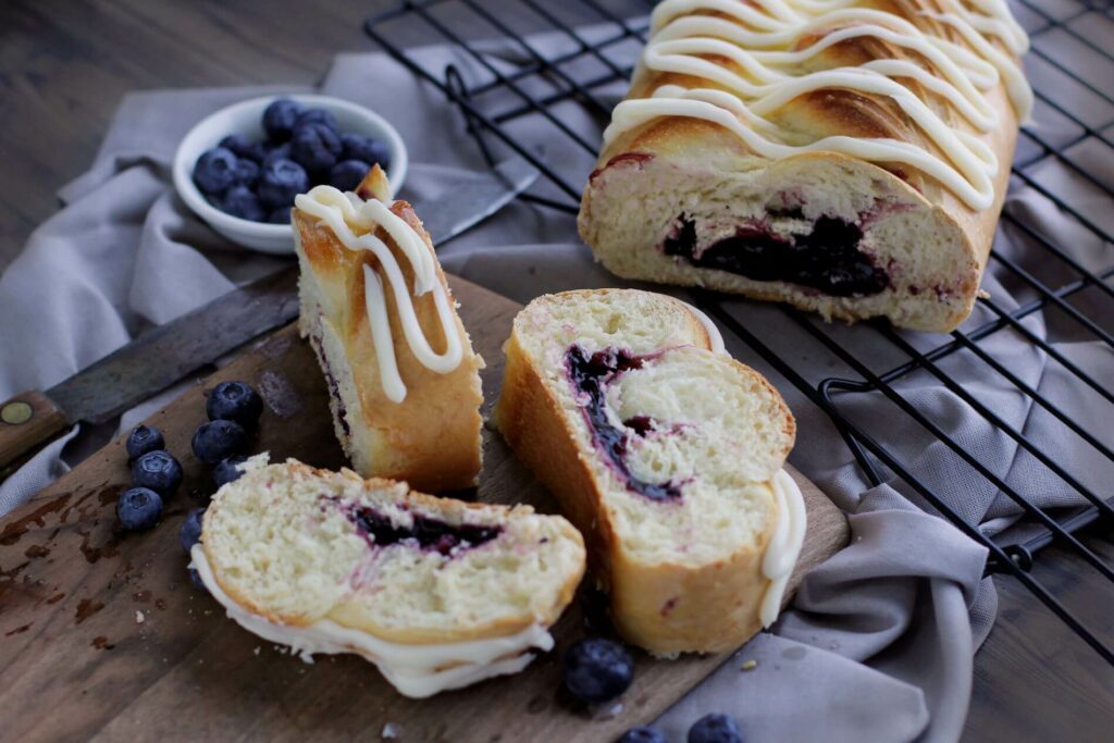 A partial Blueberry Braided Bread on a cooling rack. In front of the rack is a cutting board with three slices of the bread on it. The full bread and slices are iced. There's also a bowl of blueberries next to the slices, and blueberries scattered on the cutting board.