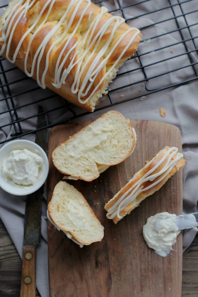 A partial Cream Cheese Braided Bread on a cooling rack. In front of the rack is a cutting board with three slices of the bread on it. The full bread and slices are iced. There's also a bowl of cream cheese next to the slices, and cream cheese smeared on the cutting board.