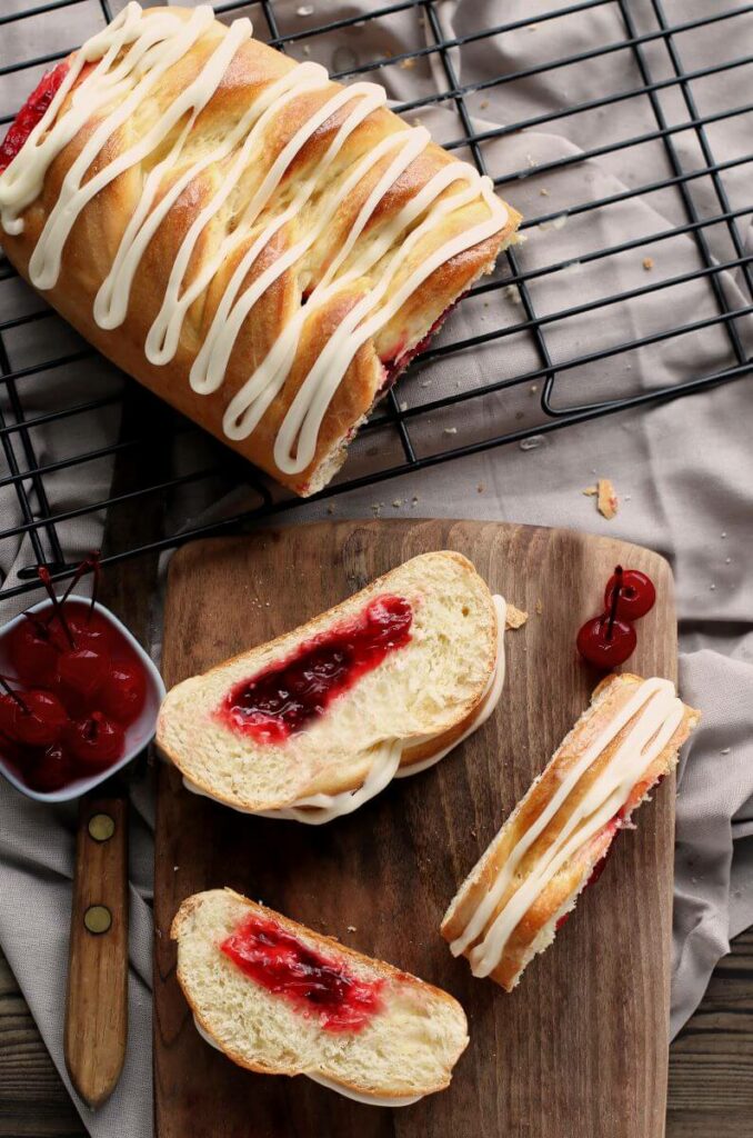 A partial Cherry Braided Bread on a cooling rack. In front of the rack is a cutting board with three slices of the bread on it. The full bread and slices are iced. There's also a bowl of cherries next to the slices.