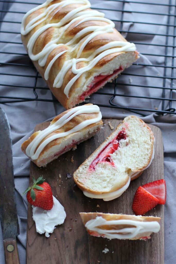 A partial Strawberry & Cream Cheese Braided Bread on a cooling rack. In front of the rack is a cutting board with three slices of the bread on it. The full bread and slices are iced. There's also strawberries, full and cut, scattered around, and cream cheese smeared on the cutting board.